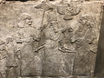 Ashurnasirapal II in the center with weapons meeting a high official after winnint the battle. An attendant protects him with an umbrella while his winged god hovers over the greeting.